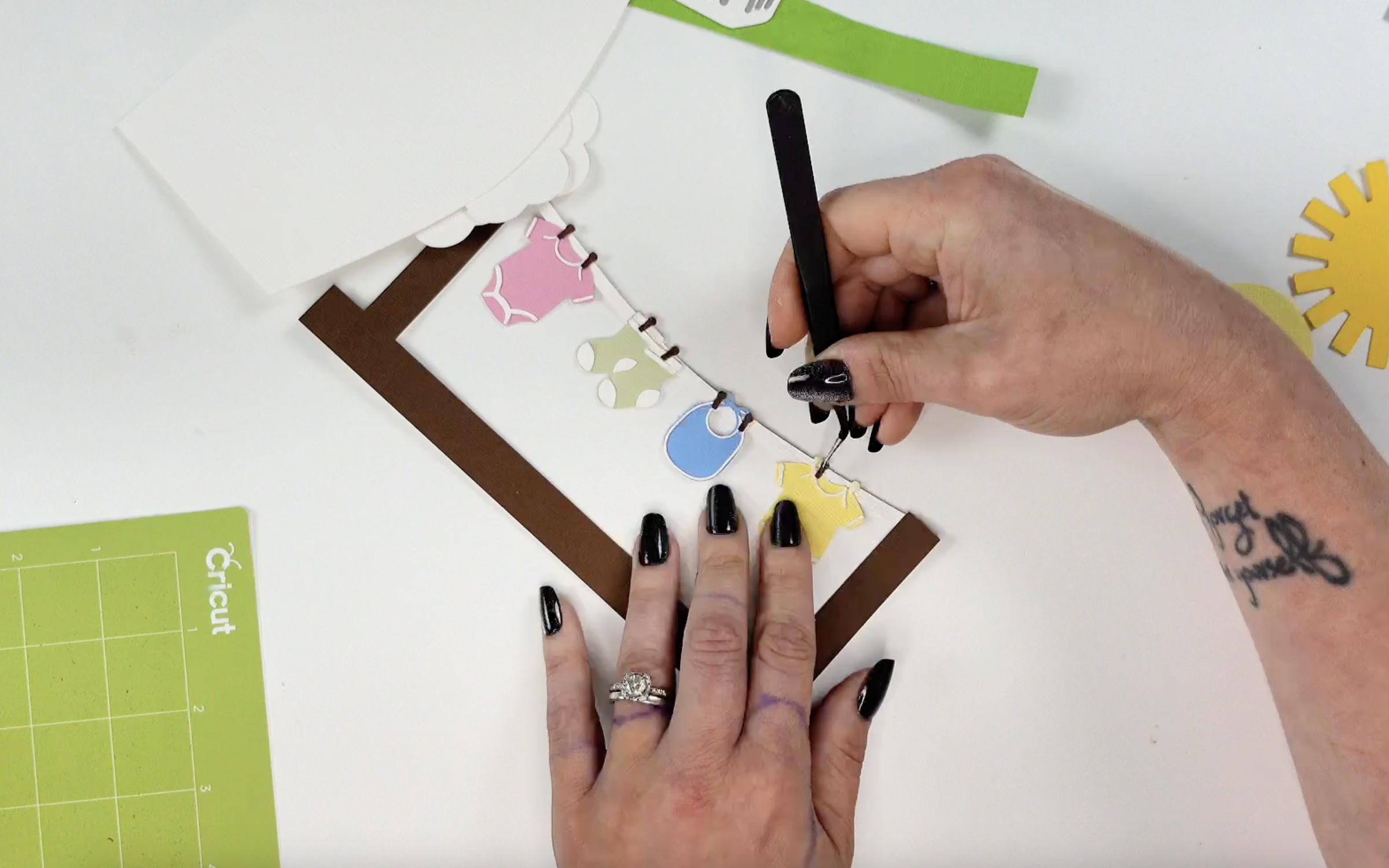 Hands using tweezers to attach small paper baby clothes to a faux clothesline on a brown cardstock frame. Pink, green, blue, and yellow baby garments are clipped to the string with mini paper clothespins.